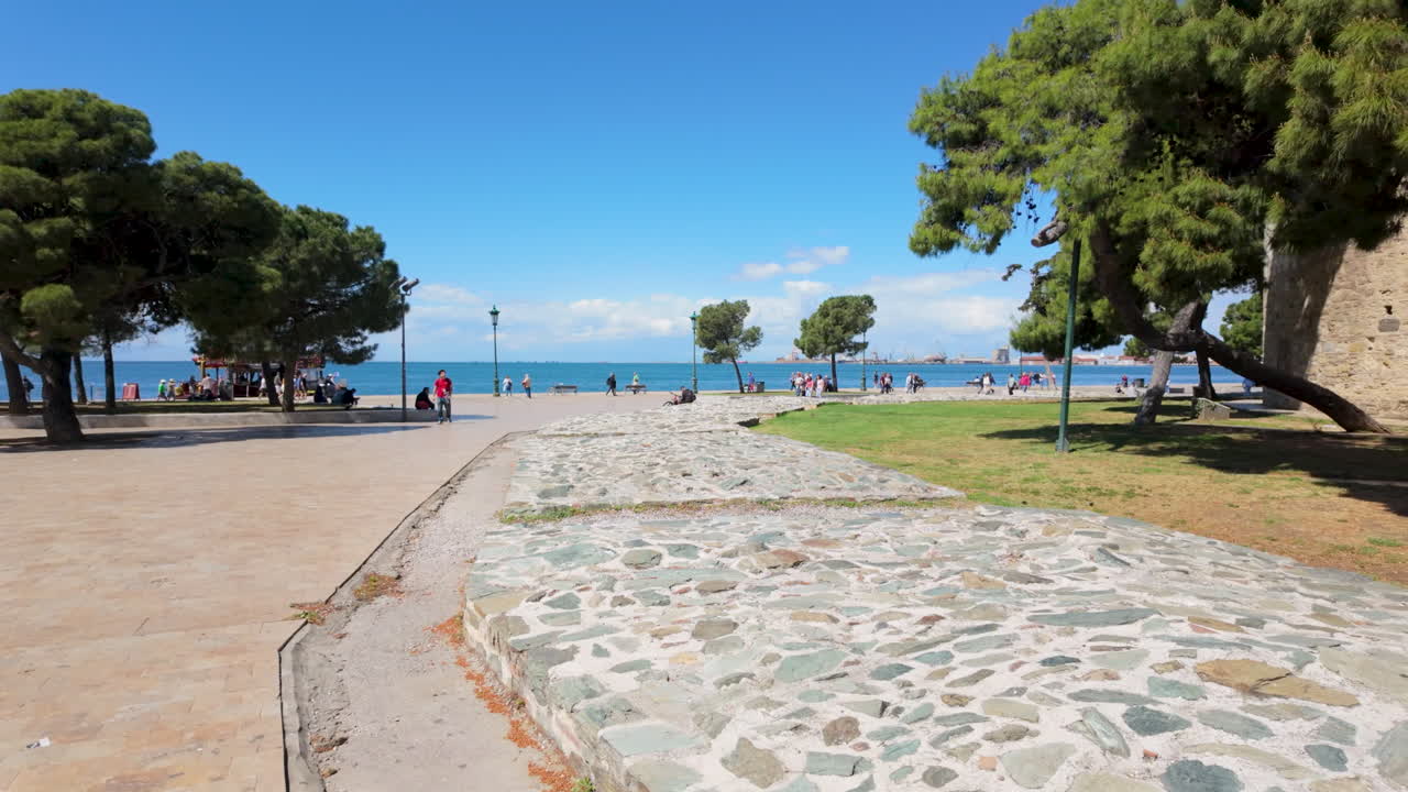 Menschen genießen einen sonnigen Tag bei einem Spaziergang durch einen malerischen Park am Meer mit Bäumen und Steinwegen in der Nähe des blauen Meeres in Griechenland
