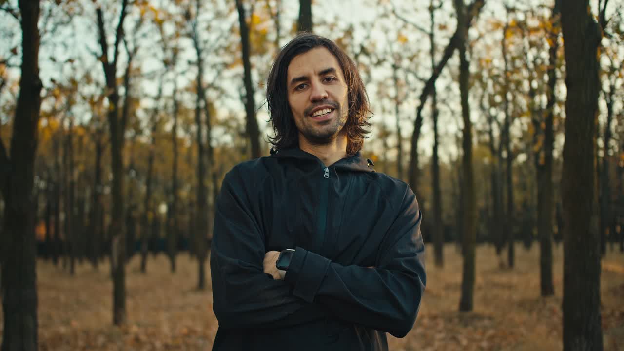 Cheerful and happy smiling man with medium length hair and a brunette beard in a black sports uniform posing in the autumn forest