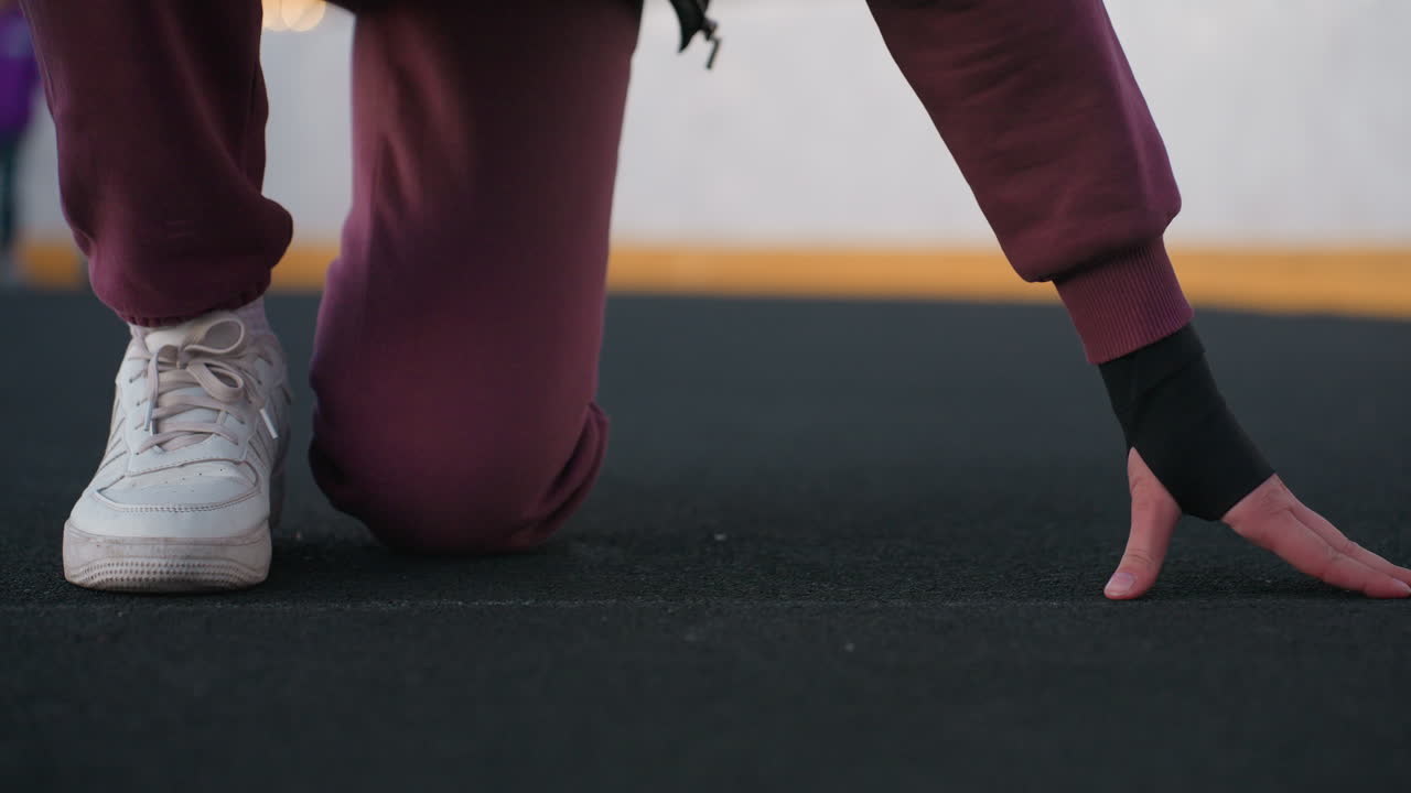 Lower view of female figure placing hands on floor with one knee on ground on black asphalt sports court near white barrier topped with chain link fence wearing maroon hoodie white sneakers