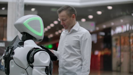 A-man-stands-with-a-robot-bot-and-asks-him-questions-and-asks-for-help-by-clicking-on-the-screen-on-the-robot-body.-Human-robot-interaction-in-the-modern-world