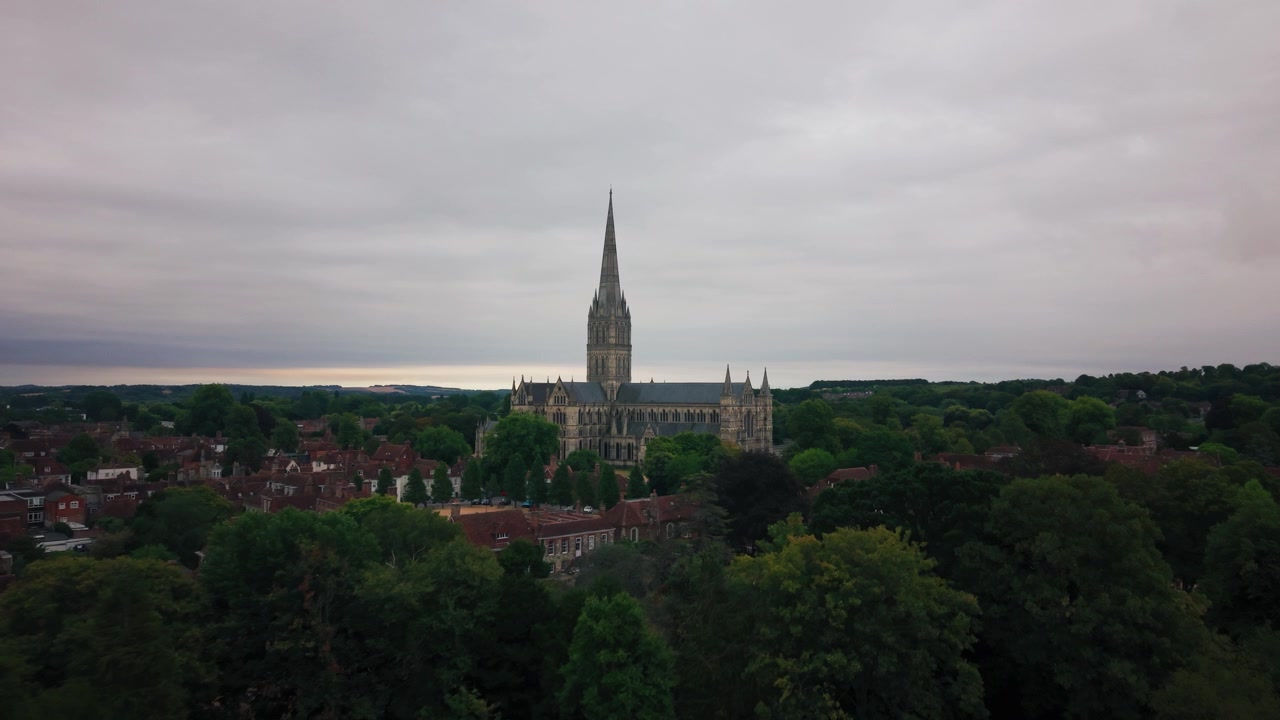 Vista aerea distante della Cattedrale di Salisbury e della sua guglia medievale che si erge sopra fitti alberi verdi sotto un cielo drammaticamente nuvoloso.