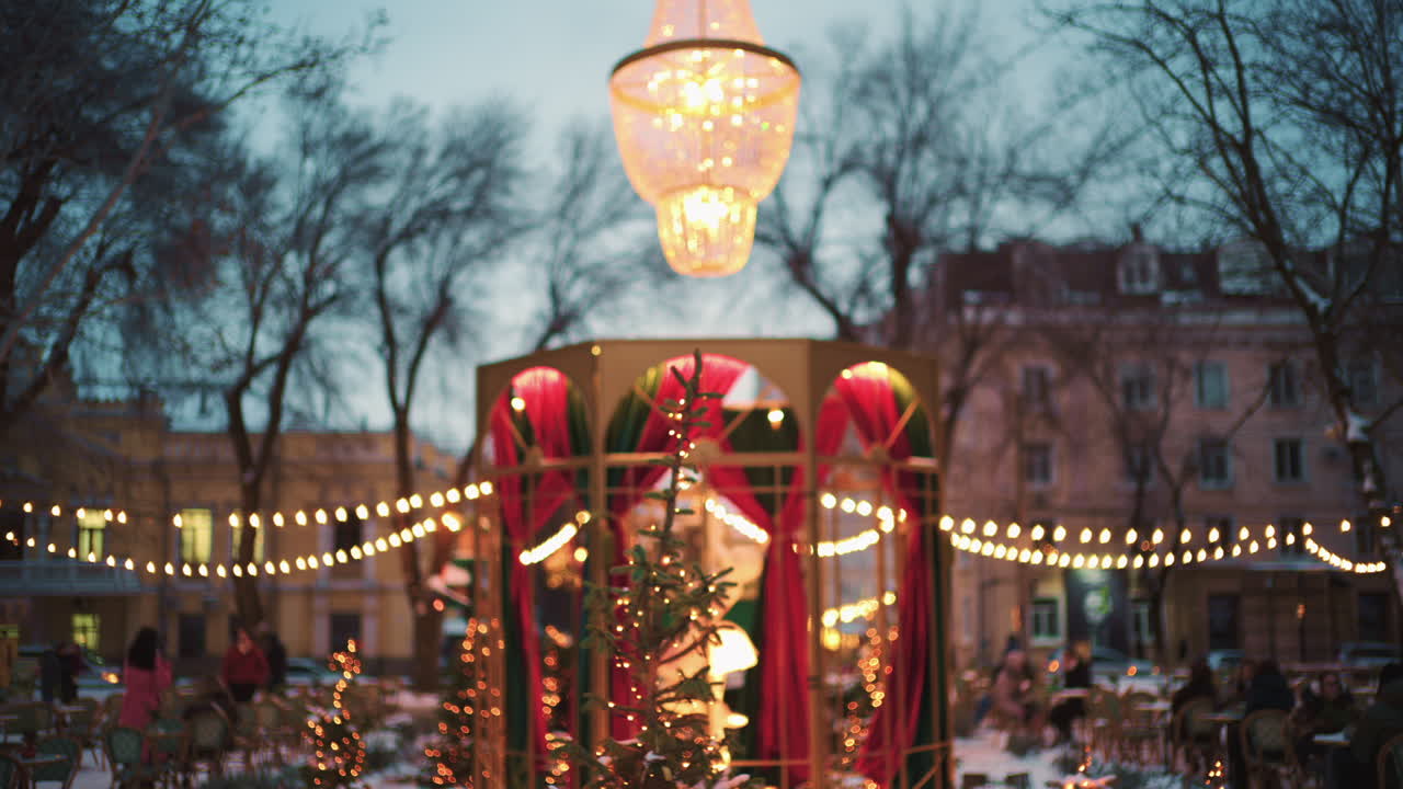 Vidvinkelbilde av festlig julepynt med lys på en utendørskafé, nær små trær og ornamenter.