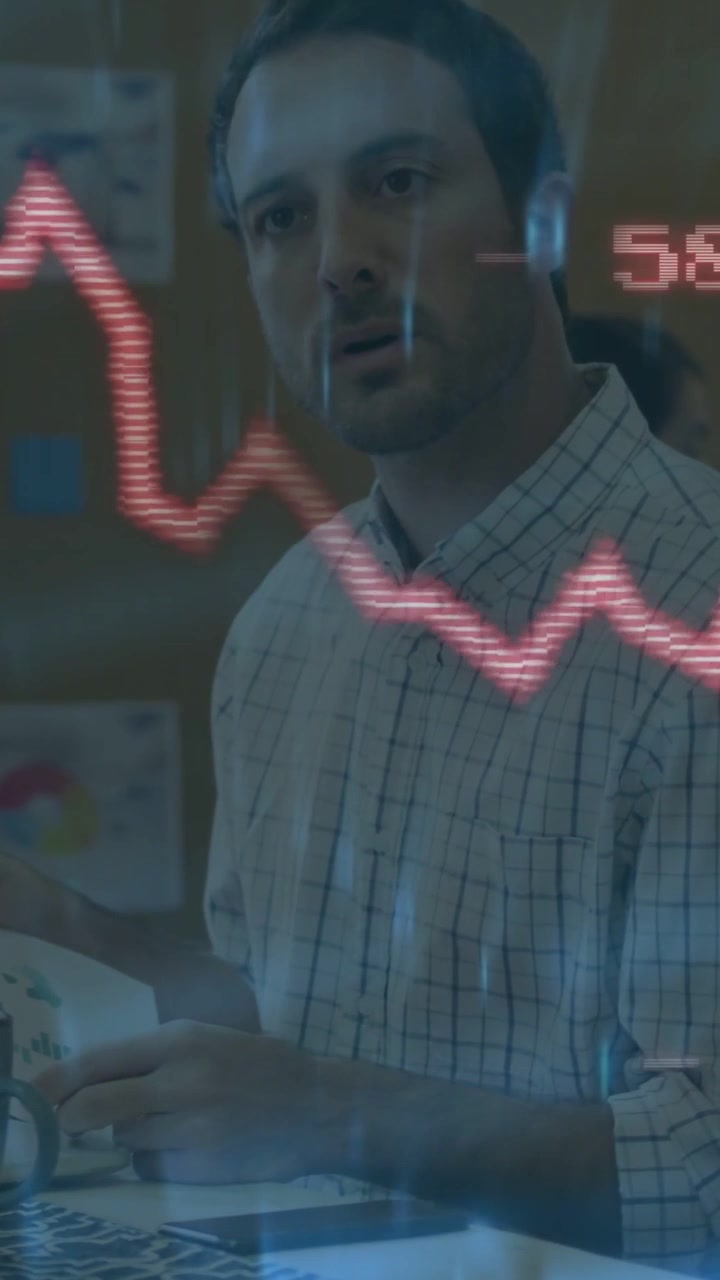 Responding to pulsing red graph, man in checked shirt at desk shifting papers and watching readouts