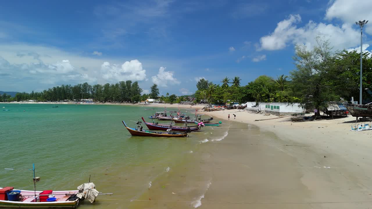Aerial flyover tracks longtail boats lining Bang Tao's calm shoreline in Phuket