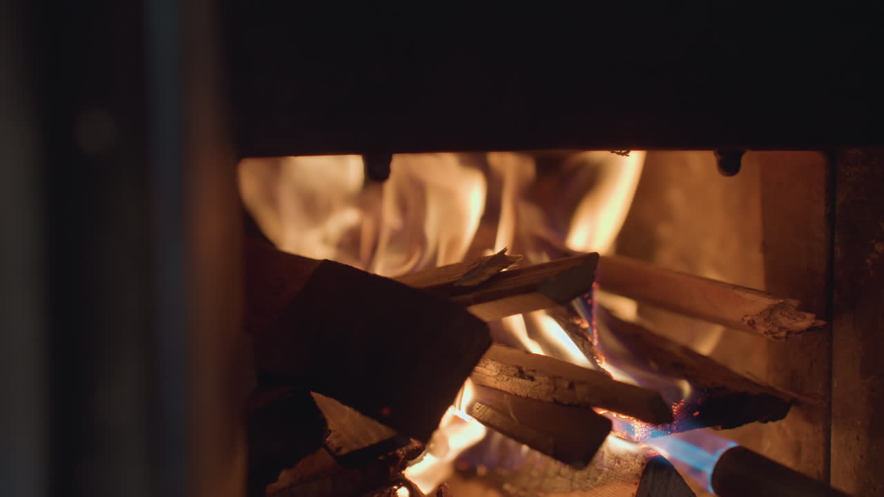 Extreme close up of individual using blue gas torch to ignite stacked firewood inside dark fireplace, flames grow gradually, illuminating wooden logs with warm flickering firelight and glowing embers