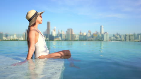 a young woman sitting in the water in an infinity-edge rooftop resort pool looks out at the city skyline of highrises