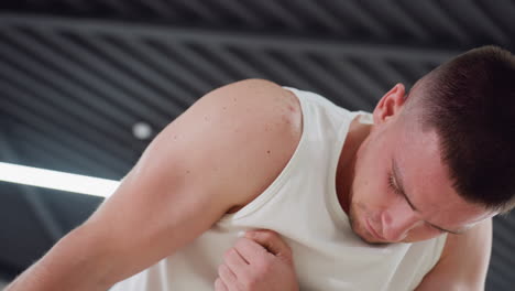 man bending head stretching arm during intense warm up move in modern fitness studio with large windows hanging straps light floor wearing white tank top black shorts and fitness watch on wrist