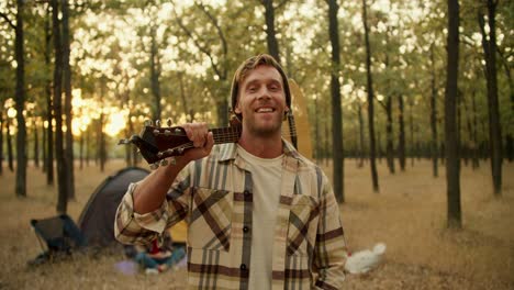 Portrait-of-a-happy-blond-man-with-stubble-in-a-checkered-shirt-who-holds-a-guitar-on-his-shoulder-while-resting-and-taking-a-break-during-a-hike-in-a-sunny-summer-green-forest