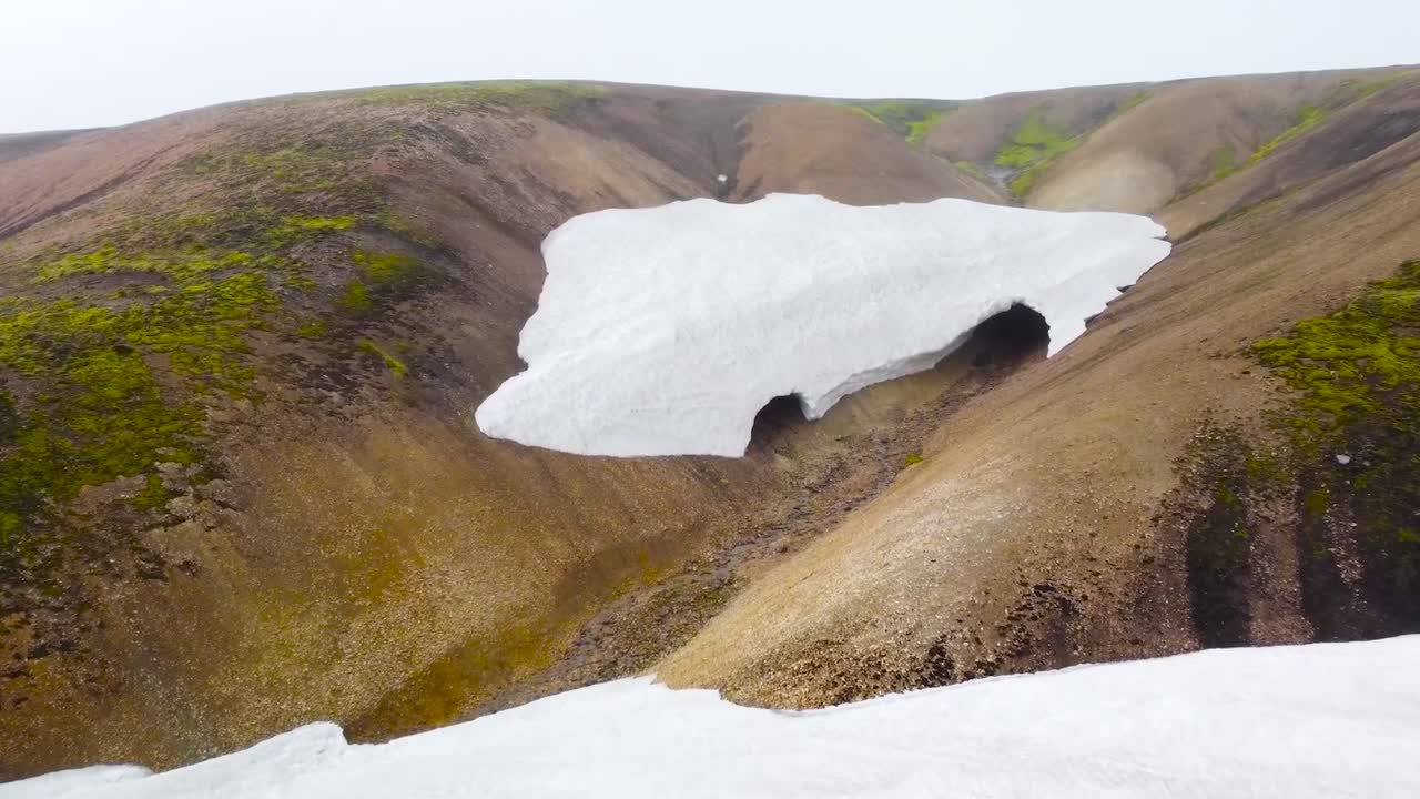Aerial footage flying closer to a mossy and snow covered Iceland slope that has some sort of cave in it. Footage gliding over glaciers or white snow covered volcanic ground during cloudy day.