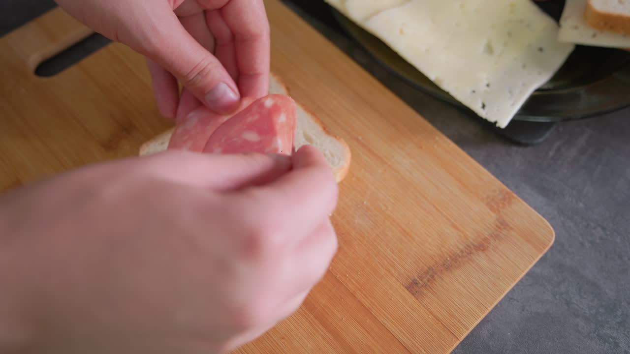 Closeup of person preparing sandwich on wooden cutting board with slice of white bread placed beside plate of cheese in home kitchen, capturing simple and routine food preparation moment indoors