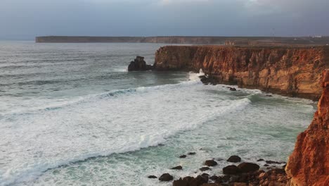 Red-sea-cliffs-of-Tonel-beach-in-south-Portugal-splashed-by-foamy-waves,-Aerial-flyover-shot