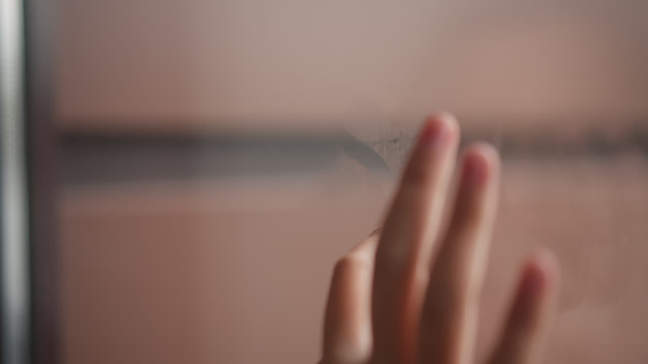 Closeup of hand and breath in cozy moment. Child playfully interacts with fogged window in indoor warmth. Tender scene of child tracing tiny shapes on fogged glass with warm breath