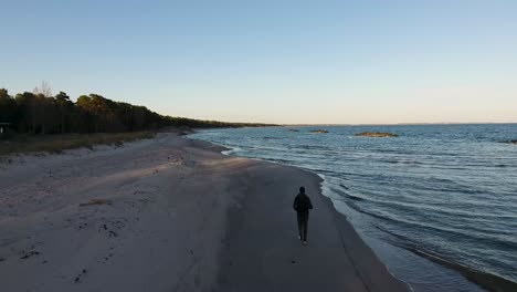 Aerial-Shot-of-a-Man-Running-By-The-Östersjö-Ocean-in-The-Evening-at-The-Ystad-Saltsjöbad-Beach-With-The-Horizon-of-South-Sweden-Skåne-in-The-Background