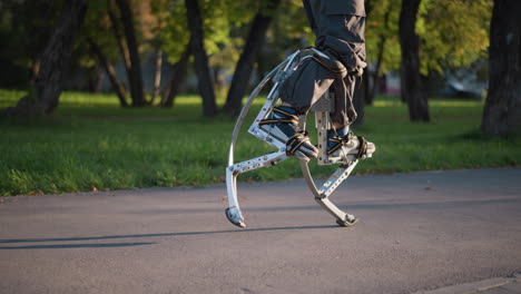 legs of man using spring stilts while walking on paved path in sunny park, wearing dark gray pants. low camera angle focuses on dynamic movement, highlighting stilts and vibrant outdoor scenery