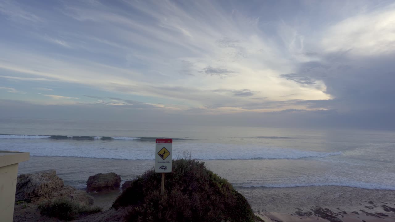 A peaceful evening scene at Point Lonsdale back beach in Victoria, Australia, showing rolling surf and a warning sign under a dramatic, wispy sky during the golden hour