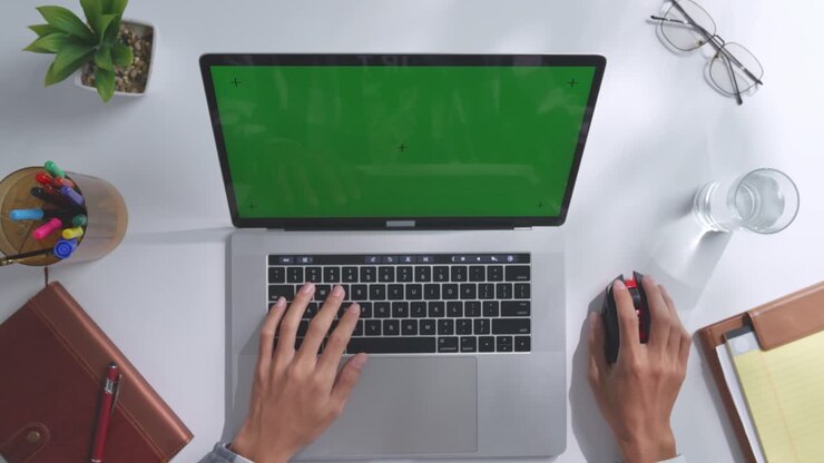 Draufsicht auf einen Laptop-Computer mit Mockup-Greenscreen-Display auf einem Holzschreibtisch, daneben ein Notizbuch mit Stiften, Brille und einem Glas Wasser. Langsames Herauszoomen, Nahaufnahme.