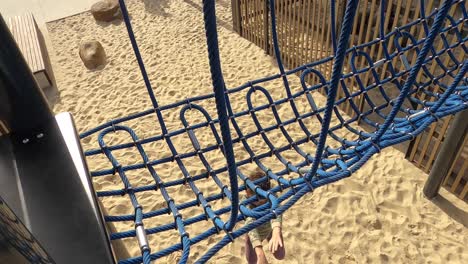 A-cheeky-little-boy-hangs-off-a-blue-rope-swing-bridge-and-pretends-to-die-or-hurt-himself-when-he-lands-in-the-sand