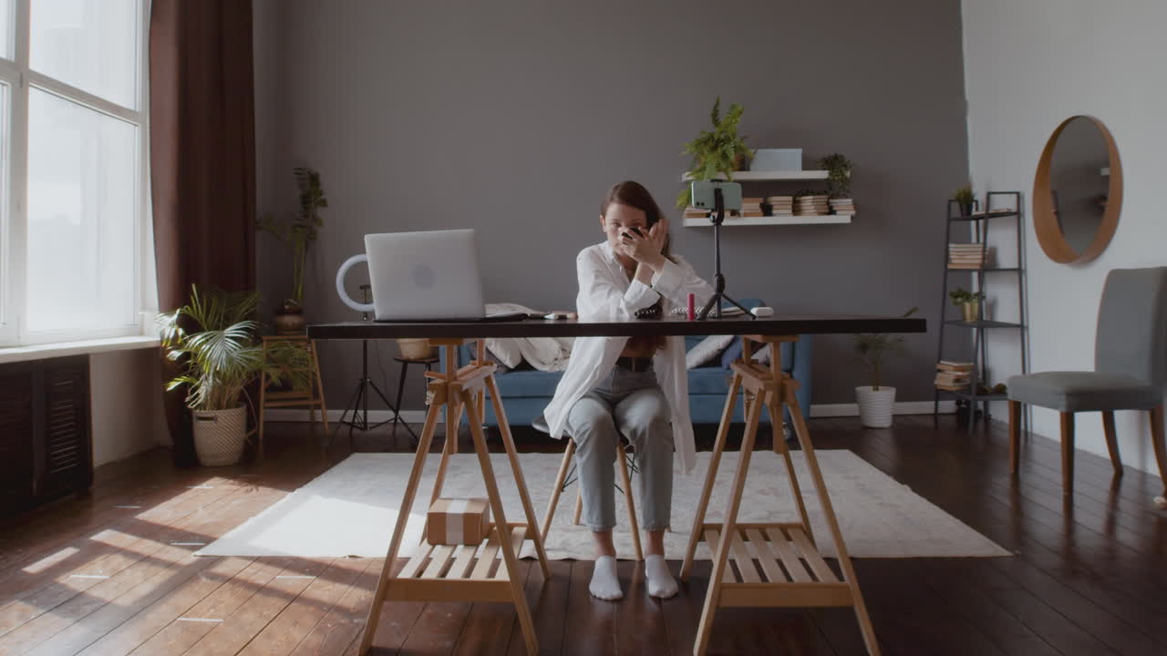 스마트폰 카메라 앞에서 눈 마스카라를 테스트하고 검토하는 젊은 동영상 블로거 여성의 와이드 샷