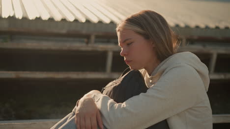 close-up of woman in deep thought, seated alone on empty stadium bleachers, hugging her bag with a reflective, introspective expression, sunlight illuminates her face