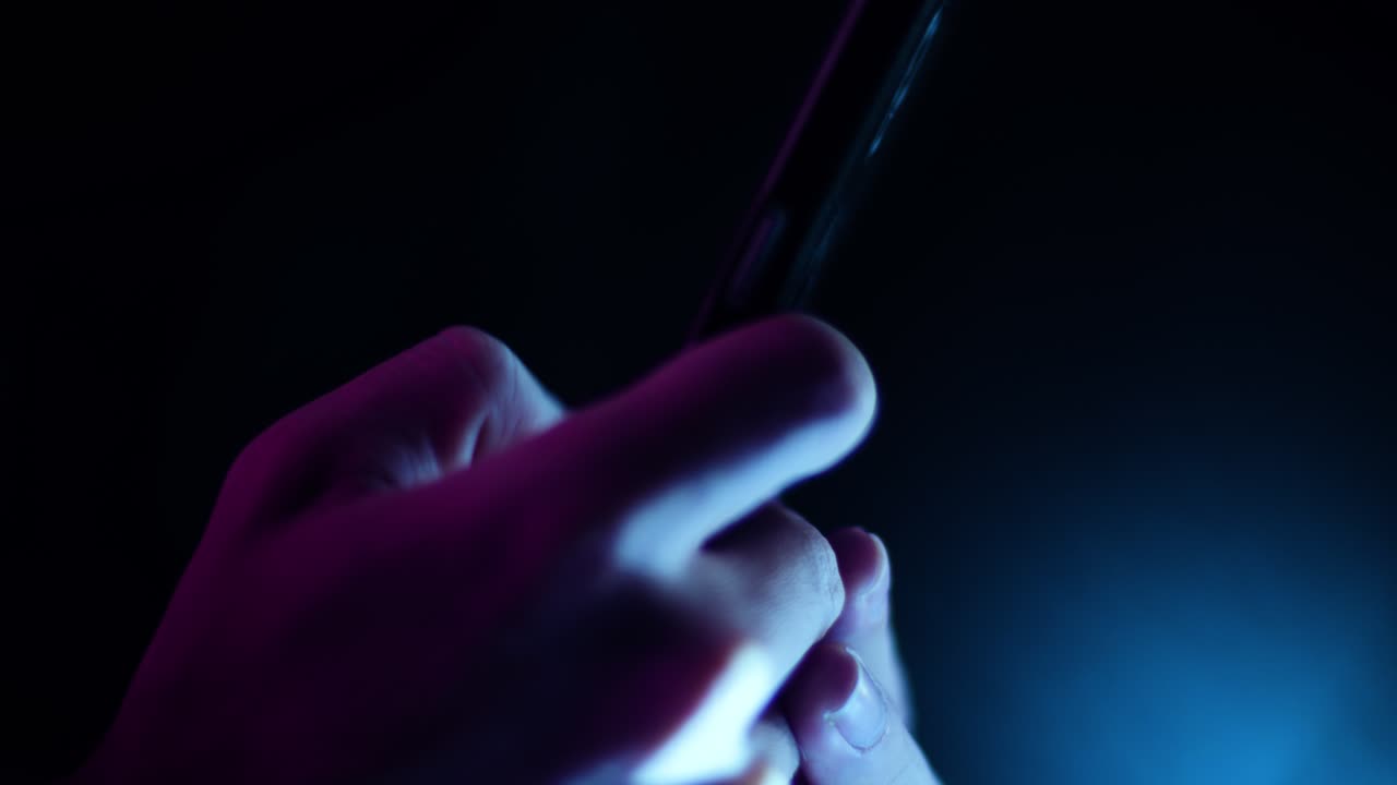 desplazándose con el pulgar y tocando un smartphone, cierre el perfil lateral en una habitación oscura con iluminación violeta y azul