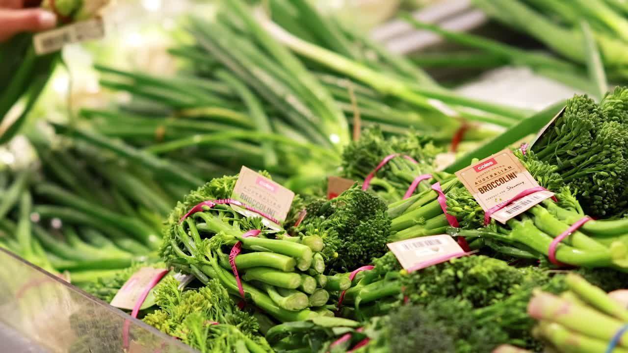 A person reaches for fresh broccoli and spring onions in a well-lit produce section.
