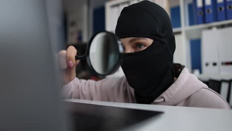 person in balaclava investigating laptop screen with magnifying glass