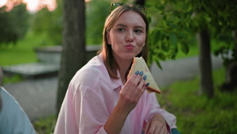 close-up of woman enjoying sandwich with a smile on her face, surrounded by green nature in a relaxed outdoor setting, with partial view of some holding sandwich