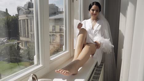 Bride-in-boudoir-dress-sitting-on-window-sill-wedding-morning-preparations-woman-in-night-gown,-veil