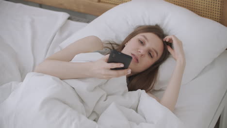 Junge-Kaukasische-Frau,-Die-Morgens-Auf-Dem-Bett-Sitzt,-Fühlt-Sich-Schlecht.-Eine-Junge-Frau-Telefoniert-Per-Smartphone-Mit-Ihrem-Arzt.