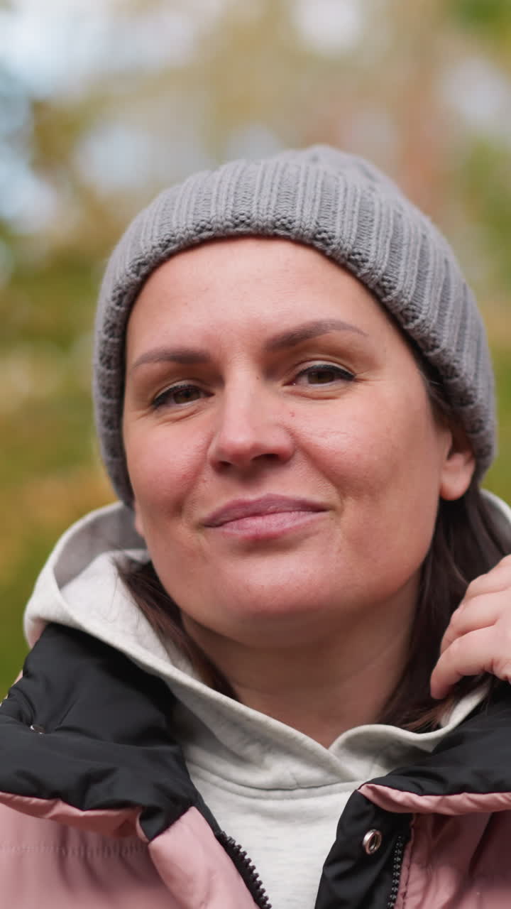 mujer sonriente con gorra de ceniza y chaqueta rosa ajustando su cuello al aire libre en otoño, rodeada de follaje borroso vibrante, expresando calidez y estilo casual con un comportamiento relajado