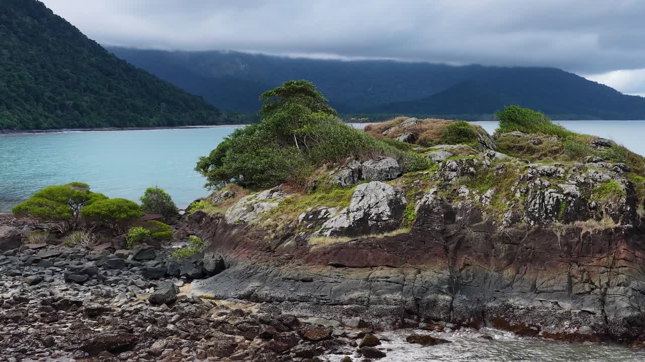 Aerial footage glides over a rocky, vegetated shoreline with turquoise waters and distant mountains under overcast skies, highlighting natural coastal scenery and dynamic camera movement