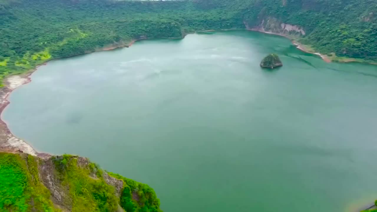 Aerial drone flying over mountains ridges that are on the edge of Taal or Bombon lake in the Philippines during a cloudy day. A large rock formation is in the blue and green water, green nature around
