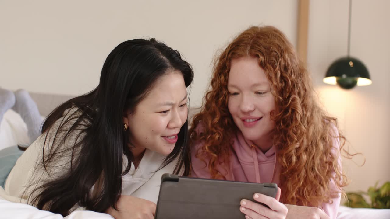 Diverse donne sdraiate sul letto, la donna a destra tiene un tablet mentre quella a sinistra si sporge per visualizzare insieme i contenuti