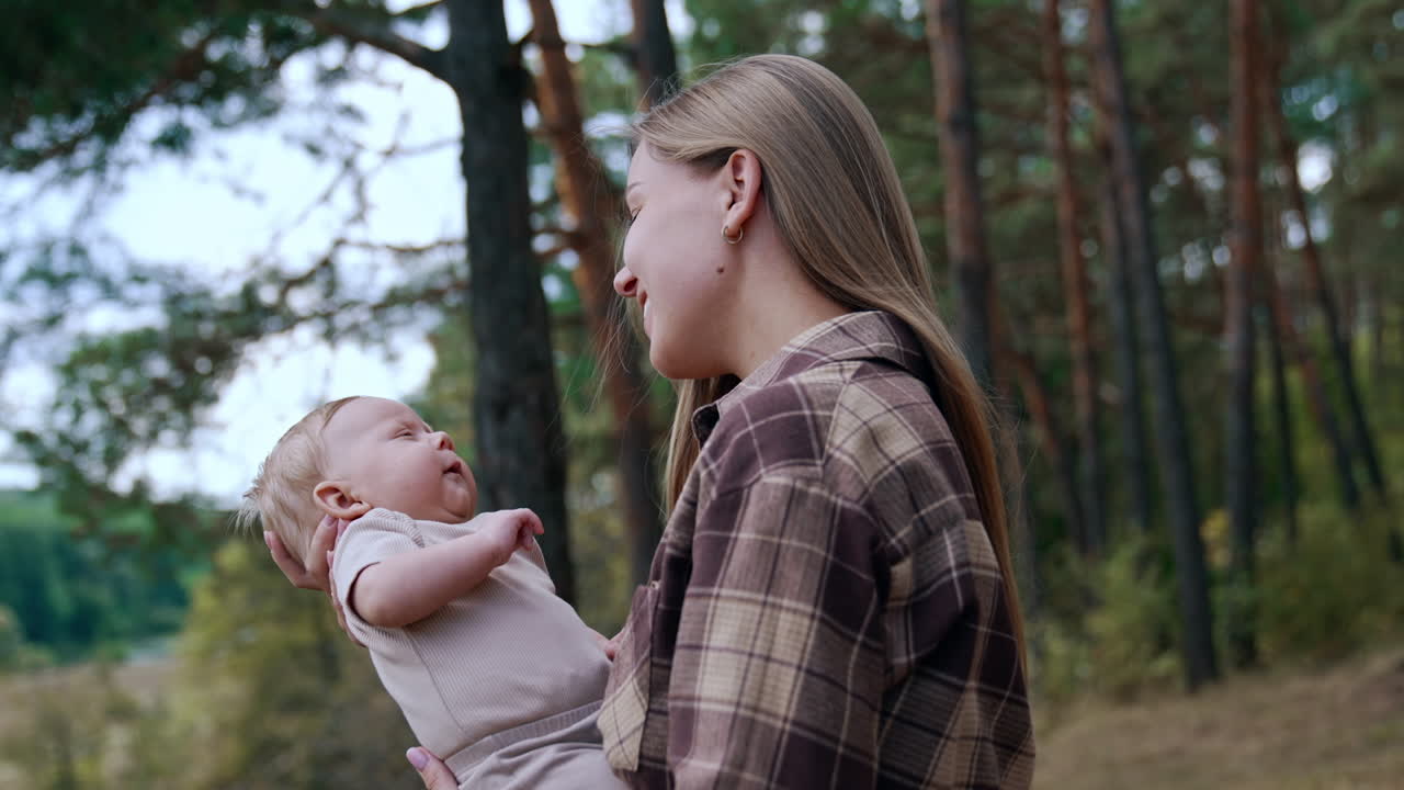 Pretty long-haired woman in checkered shirt holding her baby. Mother and infant spend time in the nature.