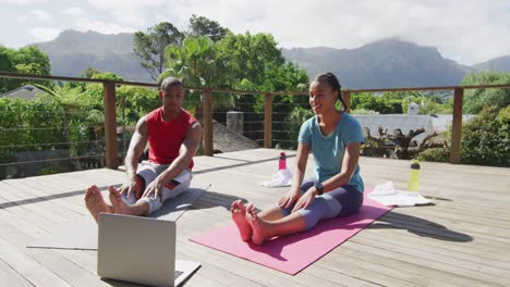 Relaxed-biracial-couple-on-terrace-exercising,-stretching-with-online-lessons