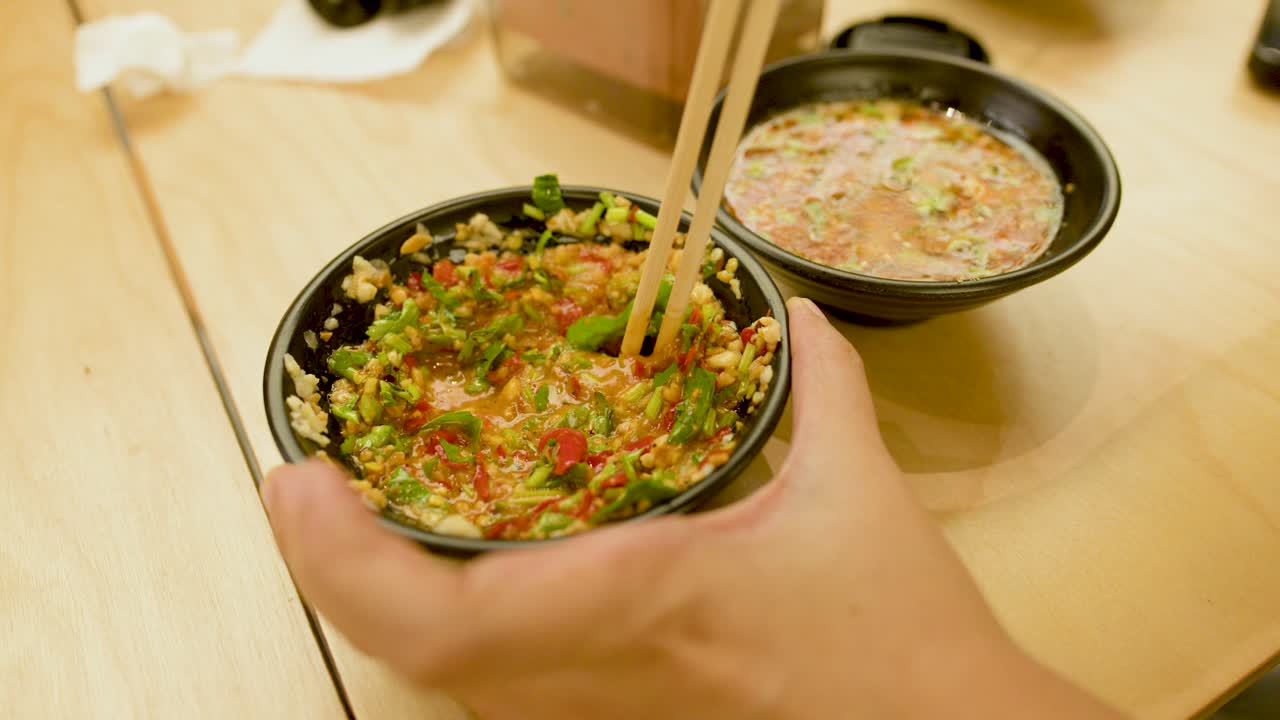 Hand uses chopsticks to mix colorful spicy rice bowl, bright lighting, wooden table, close-up