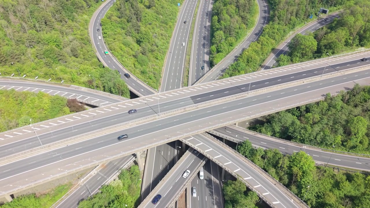 Aerial view of a sprawling highway interchange at daytime. Multiple levels of roads and bridges carry cars and trucks through a complex network. Lush green foliage surrounds the active traffic flow.