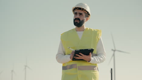 auditing wind turbines in a field of renewable energy generators on a sunny day, a professional engineer in a white helmet and reflective vest uses a tablet with technology to promote sustainability