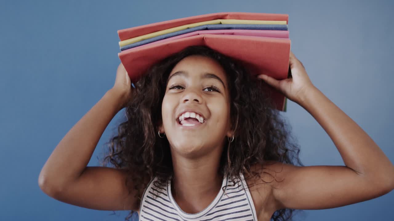 Portrait of happy african american girl holding books on head on blue background, slow motion