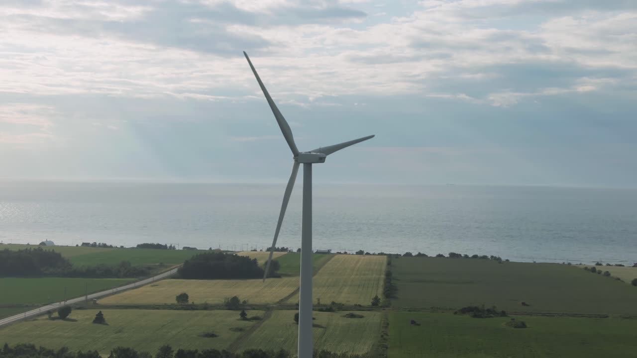 Wind Turbine Generating Clean Energy On The Green Field By The Coast Of Saint Lawrence Gulf In Northern Quebec With Sun Beams In The Background - aerial drone, panning shot