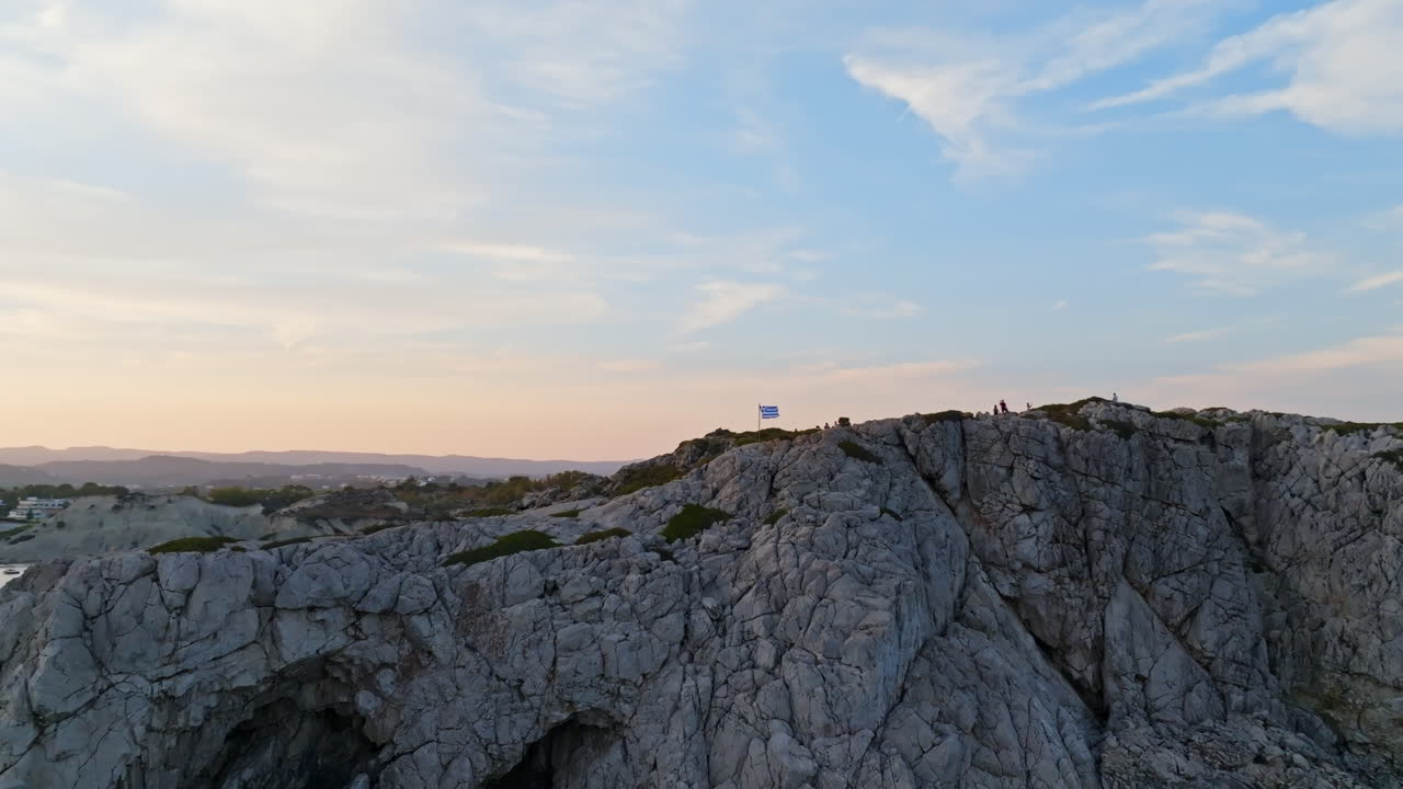 Panoramic aerial around a Greece flag on a cliff, in Kolympia, Rhodes, sunset