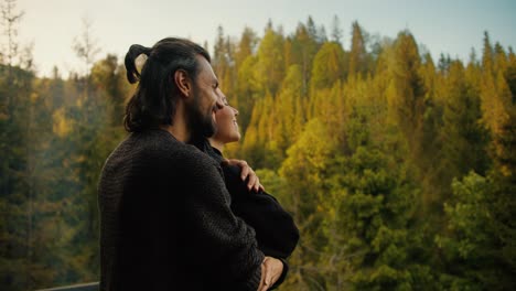 a brunette man approaches a girl on the balcony of a country house, hugs her and together they look at the mountains covered with coniferous forest