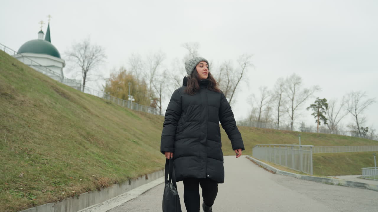 Dame spaziert mit Mantel und Mütze. Eine Frau geht zügig auf einem gepflasterten Gehweg entlang, mit Blick auf eine Kirchenkuppel. Eine warm gekleidete Frau in schwarzem Daunenmantel und Strickmütze marschiert bergauf, vorbei an einer Kirchenkuppel.