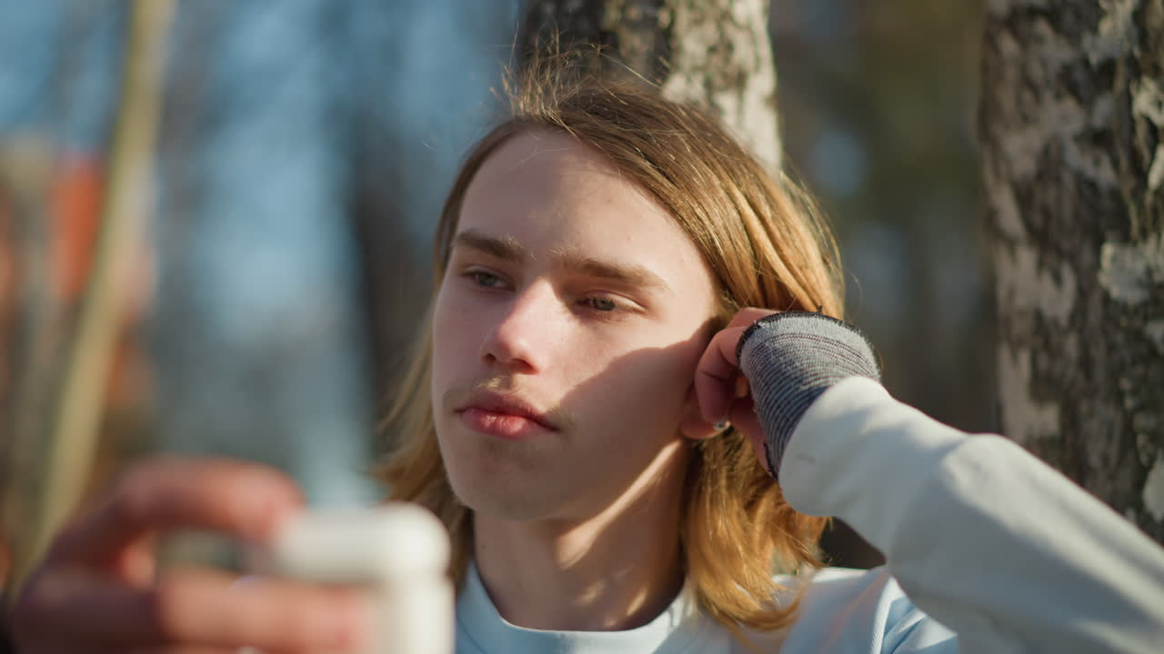 야외에서 오디오 장치를 조율하는 젊은이, 공원의 자작나무 근처에 이어폰을 꽂는 생각에 잠긴 젊은이, 차분한 태도로 야외 청취 경험 전에 오디오를 개인 설정하는 젊은이