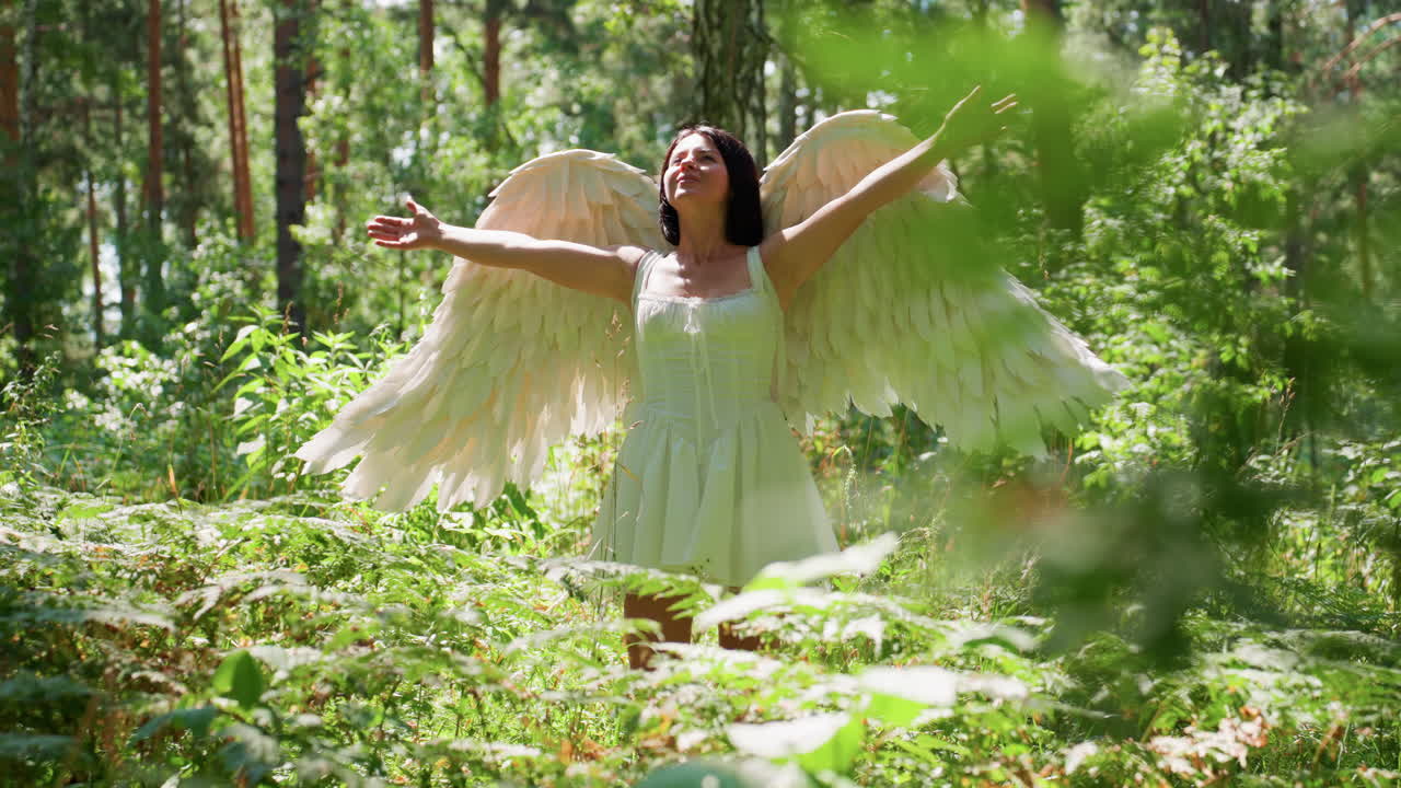 Bambina felice vestita con un costume da angelo per Halloween che allarga le braccia e si gira gioiosamente sotto la calda luce del sole in una vivace foresta verde, con ali bianche che brillano delicatamente mentre le foglie luccicano