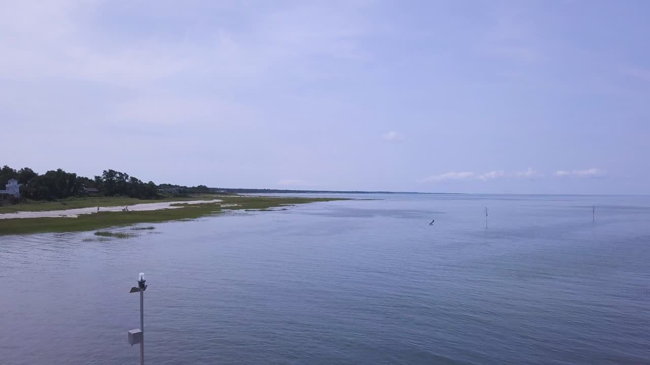 The camera pushes in and flies over the breakwater. You can see the vast expanse of the bay. Seabirds can be seen flying through the frame, and people are walking the shoreline.