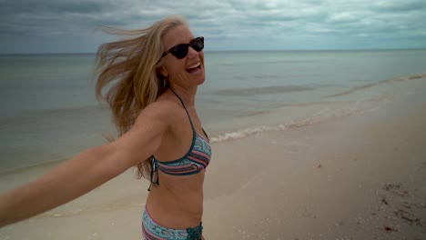 Slow-motion-with-flying-hair,-a-playful,-mature-woman-in-sunglasses-and-bikini-running-along-the-beach-with-arm-back-for-follow-me