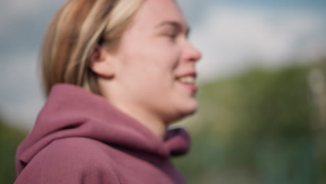 primer plano de una mujer corriendo con una sudadera roja con la capucha con el viento arruinando su cabello, el fondo borroso con vegetación y una cancha deportiva visible, capturando su movimiento enfocado y enérgico