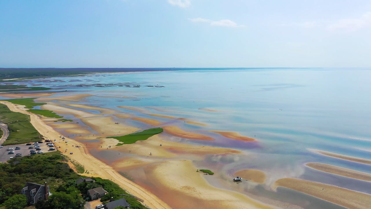 Scenic Cape Cod public beach captured by drone at ebb tide, where tidal pools shine across the sand and a parking lot filled with cars connects visitors directly to the natural shoreline.