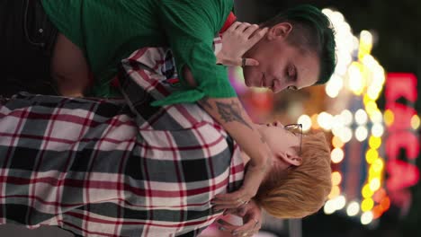 Vertical-video:-A-blonde-girl-with-short-hair-in-a-plaid-shirt-and-a-girl-with-green-hair-with-short-hair-in-a-Green-shirt-hug-during-their-LGBT-date-in-a-bright-amusement-park